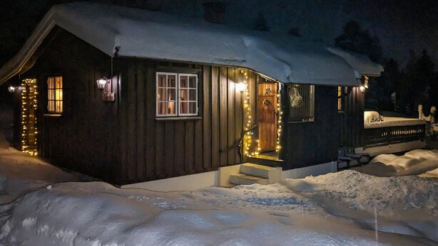 Una cabaña de madera con techo nevado y un porche iluminado con luces de adorno.