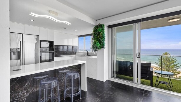 A modern kitchen with a marble countertop, bar stools, and a view of the ocean through large sliding doors. - Pacific Regis (Burleigh Heads)
