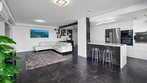 A modern kitchen with a central island, stainless steel appliances, and a seating area with a white sofa and clear stools. - Pacific Regis (Burleigh Heads)