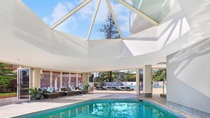 A modern indoor swimming pool with a glass ceiling, lounge seating, and a view of the surrounding landscape. - Pacific Regis (Burleigh Heads)