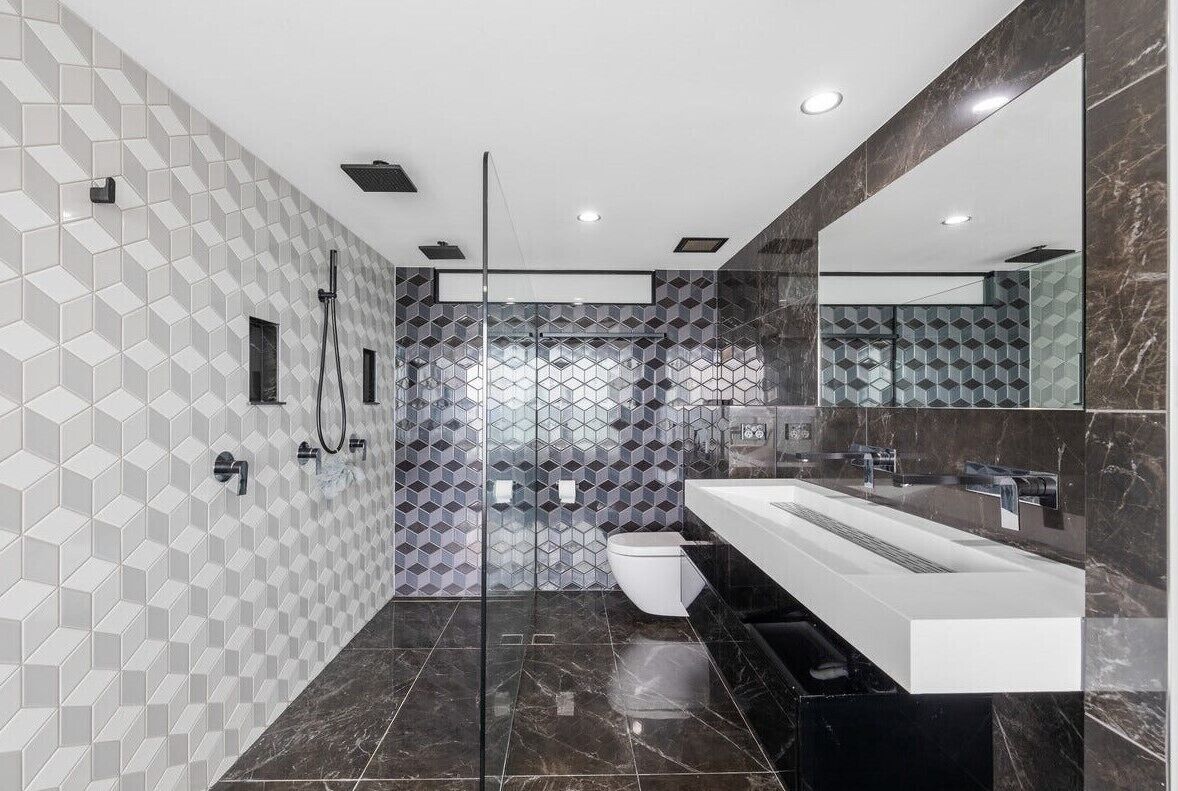 Un bagno moderno con schienale rivestito in piastrelle geometriche, uno specchio grande, un lavabo bianco e un mobile contenitore nero.