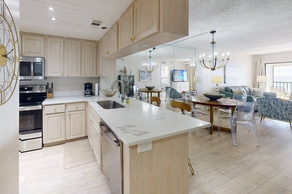 A modern kitchen with a central island, wooden cabinets, stainless steel appliances, and a dining area with clear chairs and a chandelier.