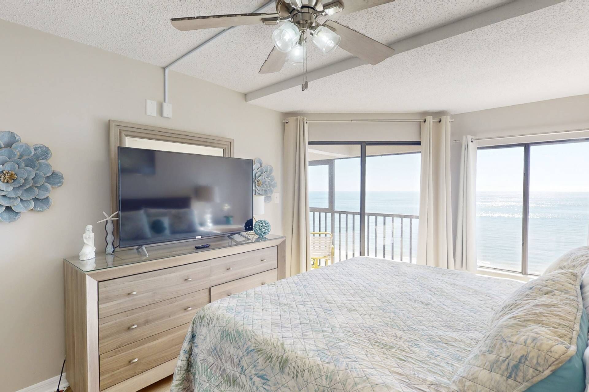 A bedroom with a large bed, a TV on a wooden dresser, a ceiling fan, and a balcony with a view of the ocean.