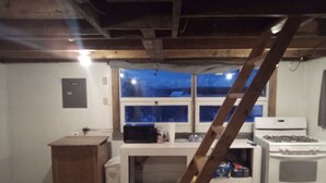 A kitchen with a stove, microwave, and cabinets. There is a window with a view of the outside. A wooden ladder is leaning against the wall. - Igloo City Cabin Rentals #1 Charming Cabin in Broad Pass at the Igloo Property (Cantwell)