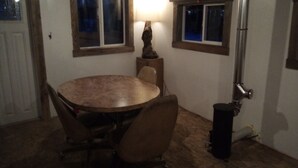 A room with a round table, four chairs, two windows with wooden frames, a lamp, and a trash can. - Igloo City Cabin Rentals #1 Charming Cabin in Broad Pass at the Igloo Property (Cantwell)