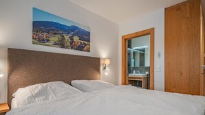 Hotellihuone, jossa on suuri sänky, puinen pääty, seinälle ripustettu maisemamaalaus, kylpyhuone peilillä ja pesualtaalla sekä puinen ovi.