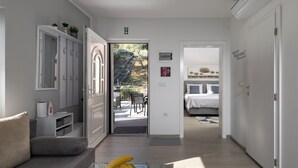 Ett modernt hotellrum med en soffa, ett matbord med en fruktskål, en garderob och utsikt över en trädgård och ett utomhusområde med sittplatser.