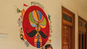 A person sitting on a wooden bench with a colorful cushion, a small table with books, and a wall with a large decorative piece. - Banana Hostel (Pushkar)