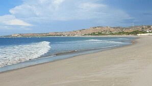 Ein Strand mit blauem Himmel, ruhigem Wasser und einer entfernten Aussicht auf Hügel und Gebäude.