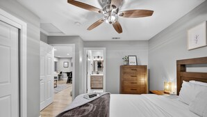 Un dormitorio con cama, cómoda de madera, mesita de noche, ventilador de techo y pasillo que conduce a un baño.