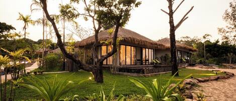 A thatched-roof bungalow with a pool, surrounded by lush greenery and palm trees.