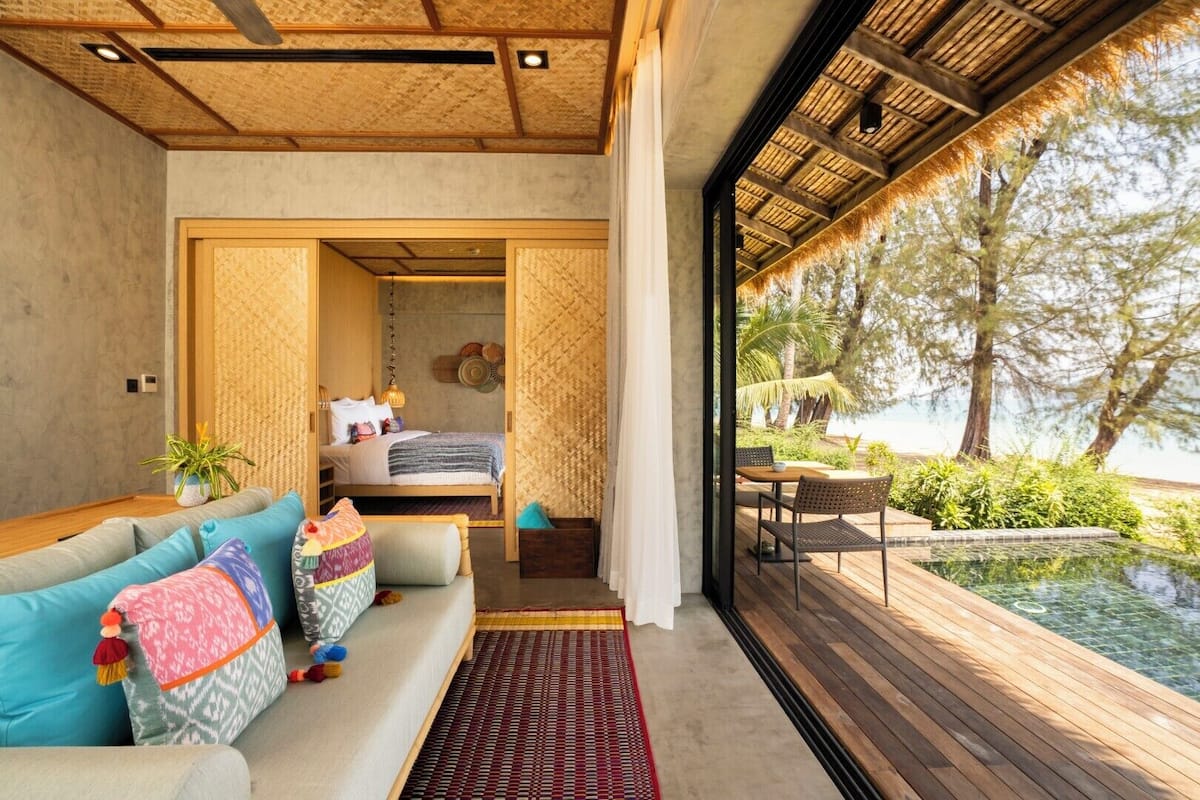 Quarto moderno com teto de madeira, cama, sofá e vista para uma piscina e vegetação lá fora.
