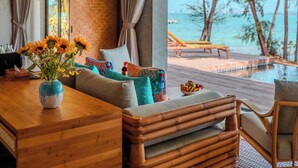 A wooden dining table with a vase of flowers, a sofa with colorful cushions, and a view of a pool and beach. - JATI Koh Russey (Prey Nob)
