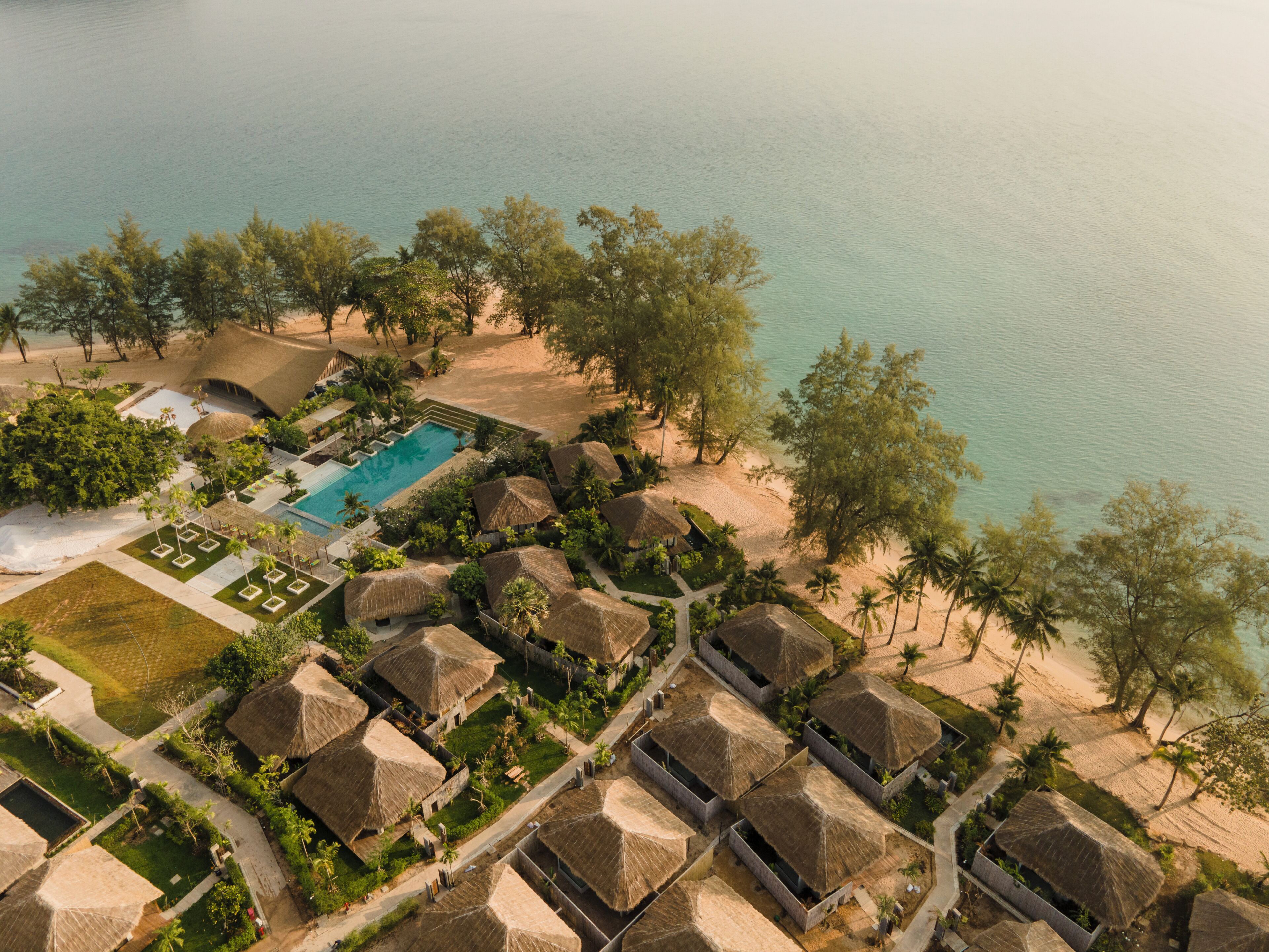 Vista aérea de un resort con bungalows de techo de paja, una piscina y una playa.