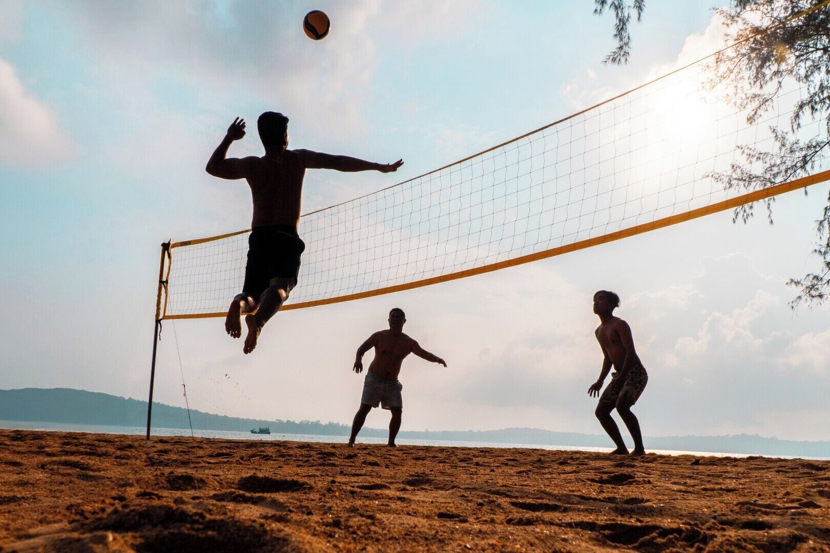 A beach volleyball game with players in shorts and no shirts, a volleyball net, and a clear sky.