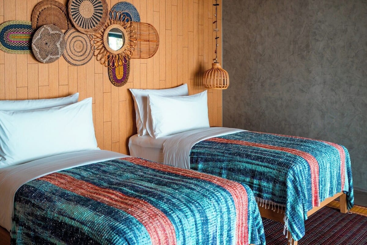 Quarto com duas camas, cabeceira de madeira, um pendão de parede tecido e luminária suspensa.