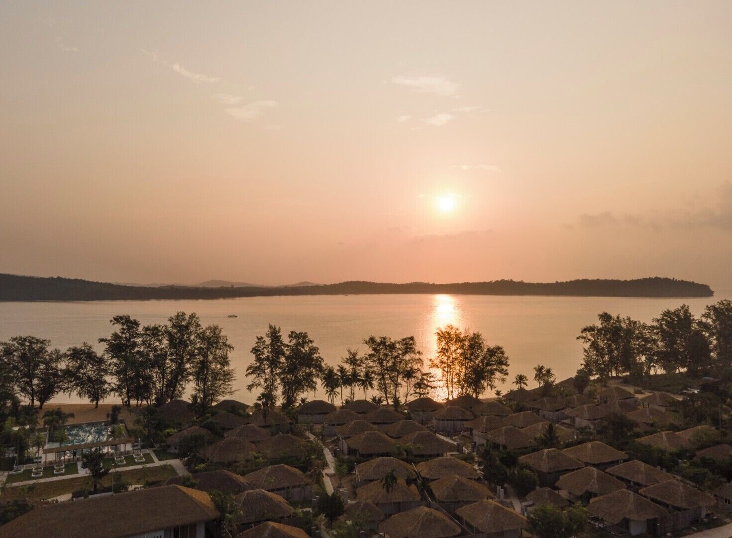 A resort with thatched-roof huts, a pool, and a view of the sunset over a calm lake.