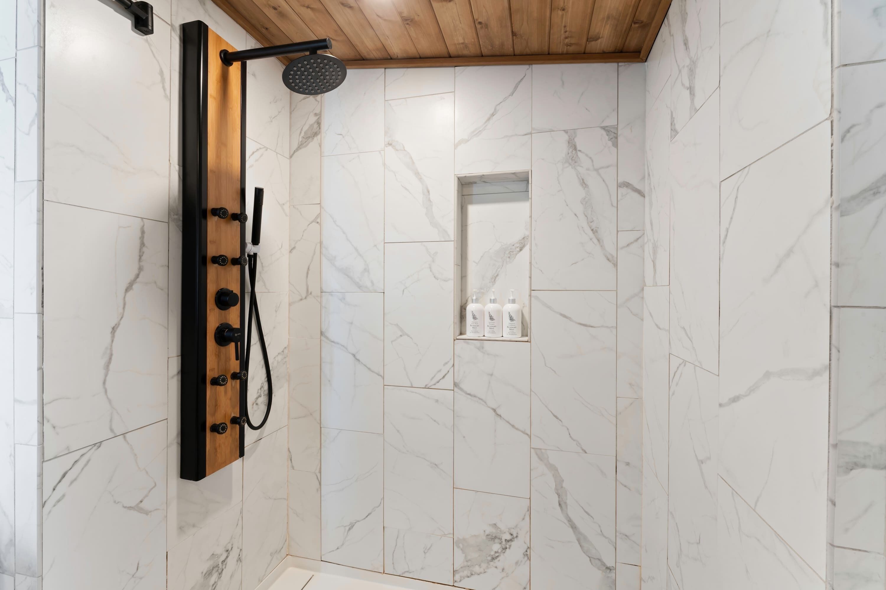 A shower with multiple nozzles and a handheld showerhead, a wooden ceiling, and marble tiles.