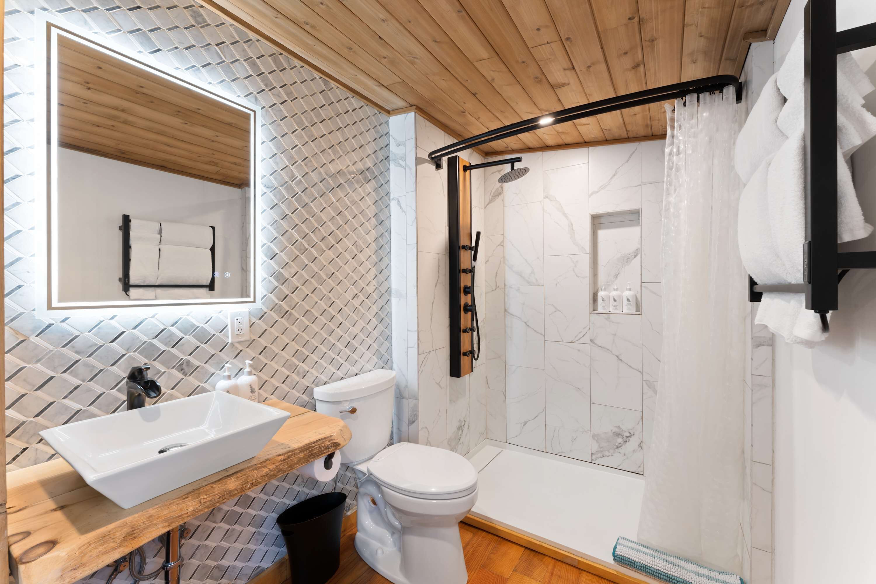 A modern bathroom with a wooden vanity, a white toilet, a shower with a black handle, and a tiled wall.