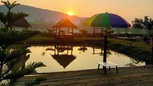 Une terrasse en bois avec une table et des chaises, un petit chalet et un parasol coloré, le tout dans un cadre extérieur paisible, au moment du coucher du soleil.