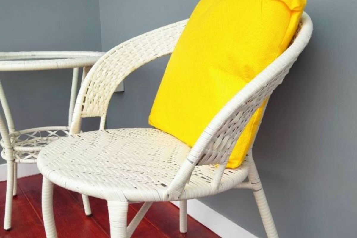 Una silla de mimbre blanco con un cojín amarillo frente a una pared gris.