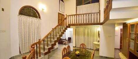 A spacious living area with a marble floor, a wooden staircase, a dining table with chairs, and a second-floor balcony with curtains.