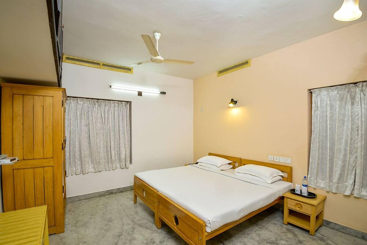 A bedroom with a single bed, a wooden wardrobe, a nightstand, a ceiling fan, and a wall-mounted light fixture.