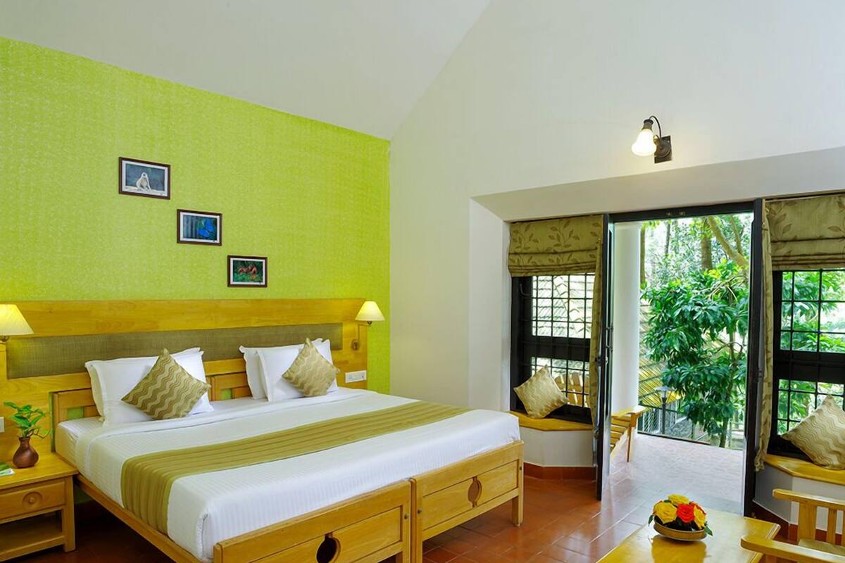 A hotel room with a large bed, wooden headboard, two bedside tables with lamps, a small table with a flower vase, and a sliding glass door leading to a balcony with a view of greenery.