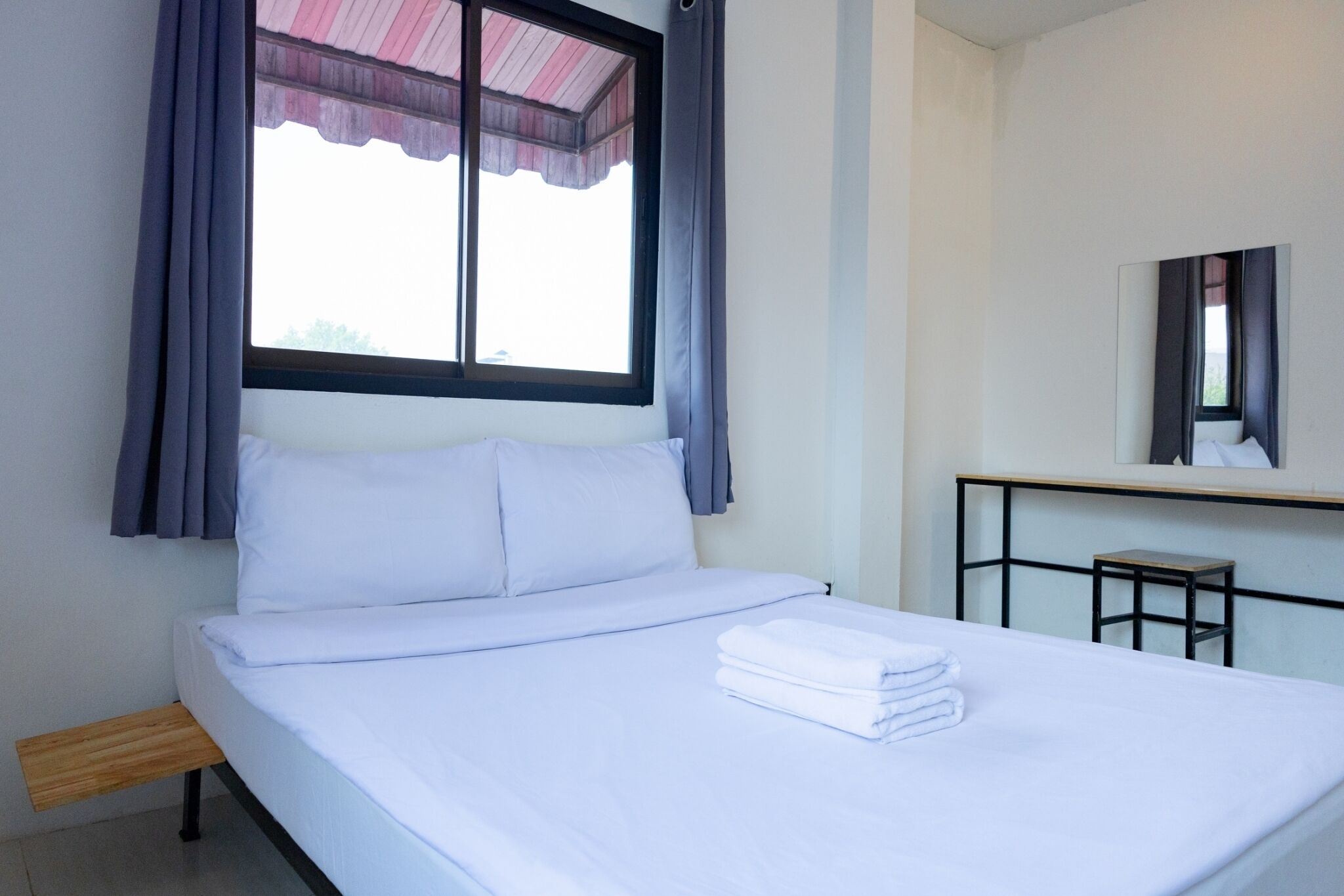 A neatly made bed with white linens and a folded towel on the side, a small wooden bench, a window with curtains, and a mirror on the wall.