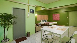 Una habitación con cama, una mesita, una silla, una planta en maceta y una puerta.