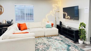Een moderne zithoek met een witte bank, een vloerkleed met dessin, een flatscreen televisie, een vloerlamp en een potplant.