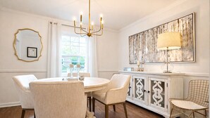A dining area with a round table, four chairs, a chandelier, a cabinet with decorative doors, and a large abstract painting on the wall. - BBQ • Swingset • Pets OK • Family Retreat (Charlotte)