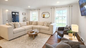 A living room with a sectional sofa, a round coffee table, and a side chair. There are two windows with curtains, a door, and a wall-mounted TV. - BBQ • Swingset • Pets OK • Family Retreat (Charlotte)