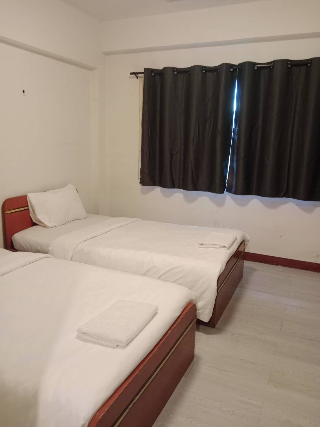 Una habitación con dos camas individuales, ropa de cama blanca, cabecera roja, ventana con cortinas negras y pared blanca.