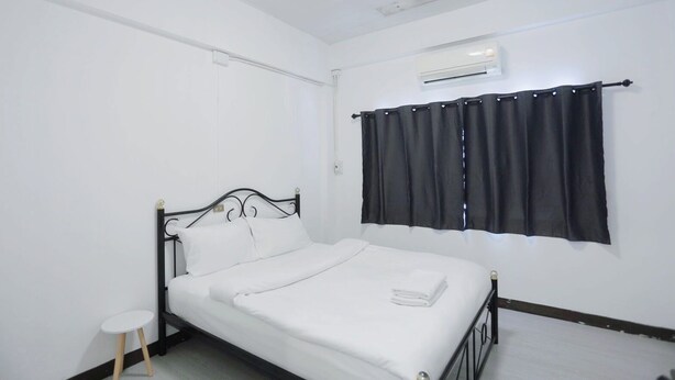 A single bed with a white mattress and black metal frame, a small white table, a black curtain, and an air conditioning unit on the wall. - GO INN Maya Chiang Mai (Chiang Mai)