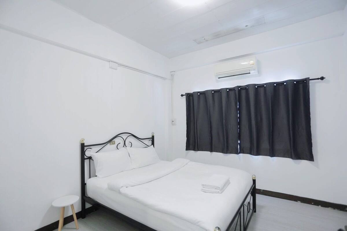 Una cama individual con colchón blanco y estructura metálica negra, una mesita blanca pequeña, una cortina negra y un equipo de aire acondicionado en la pared.