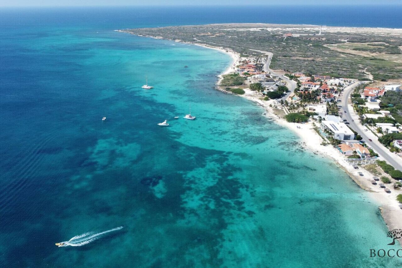 An aerial view of a coastal area with clear turquoise waters, boats, and a developed beachfront with buildings and roads.