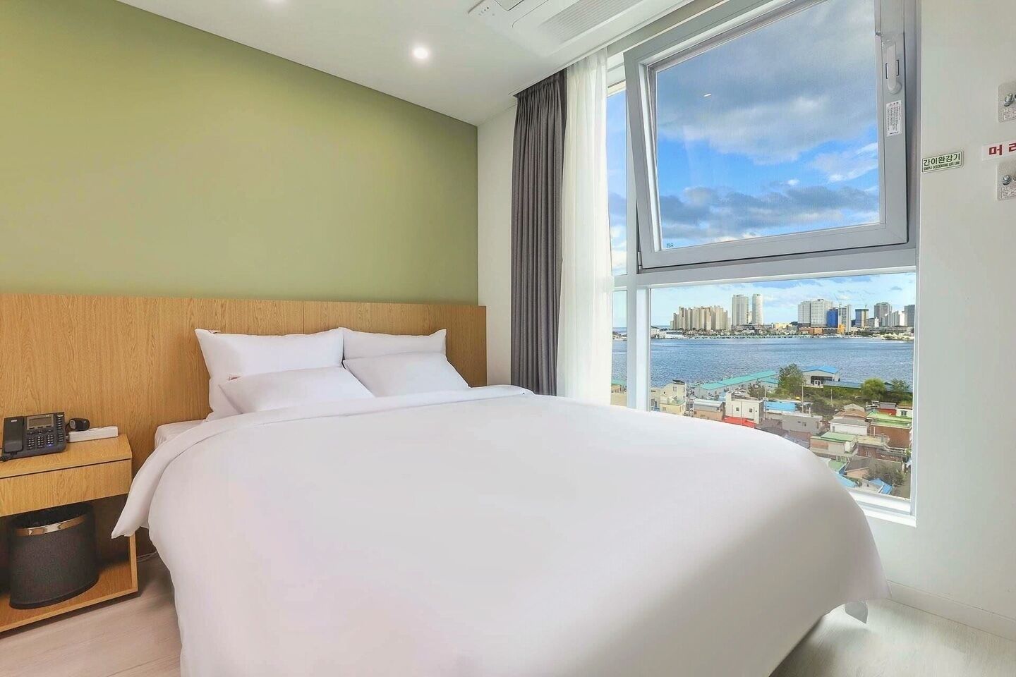 A hotel room with a large bed, a wooden headboard, a nightstand with a phone, and a window with a city view.