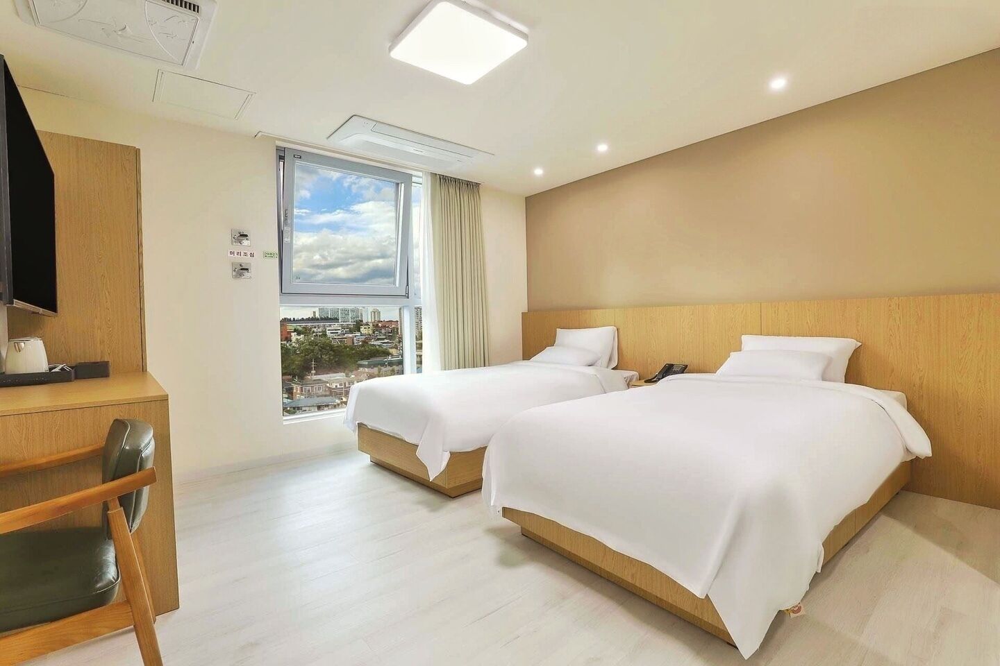 A hotel room with two beds, a desk, a chair, and a large window offering a city view.
