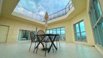 A spacious indoor area with a high ceiling, a dining table with chairs, large windows offering a view of the city, and a balcony with a railing.