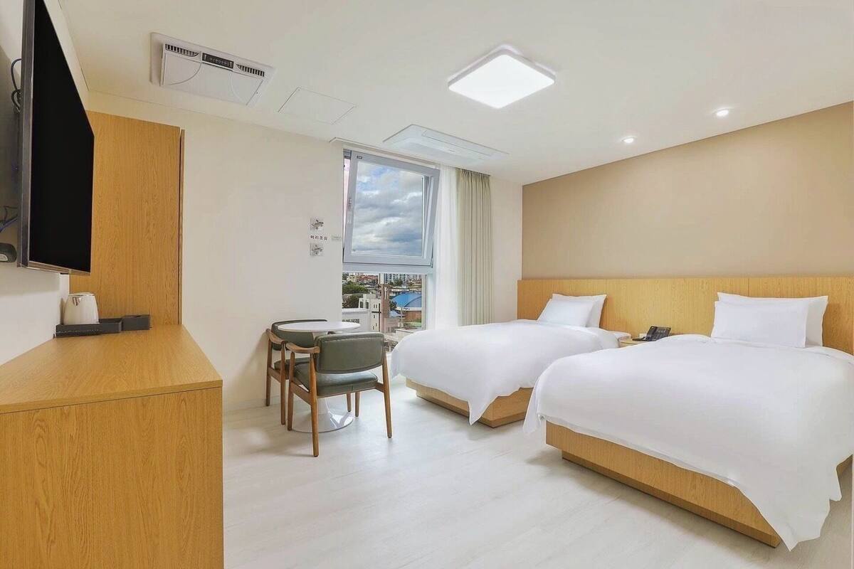 A hotel room with two beds, a desk with a chair, a TV, and a window with a city view.
