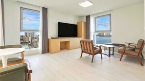 A modern living room with a flat-screen TV, wooden furniture, and large windows offering city views. - Hotel BERITH (Sokcho)