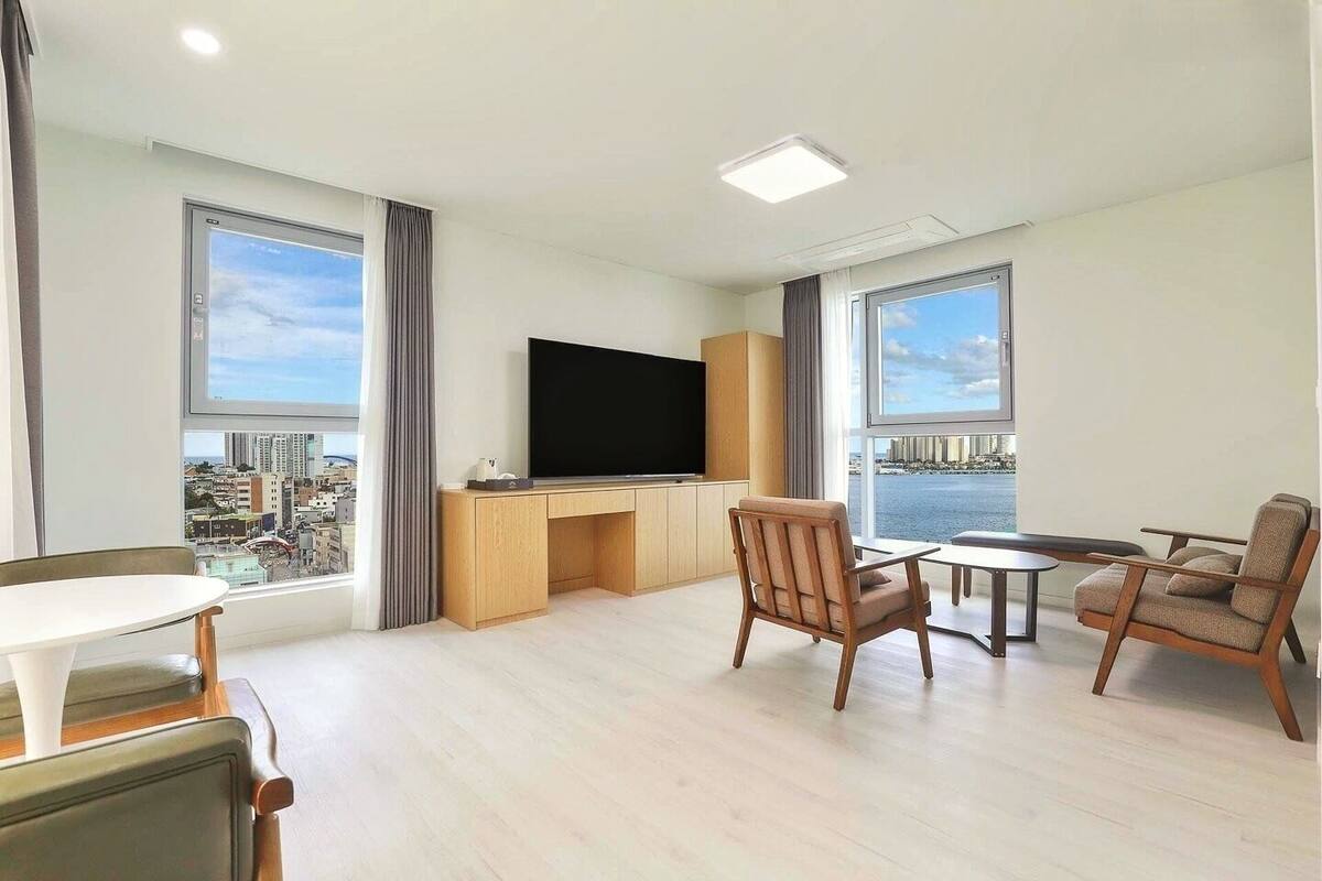 A modern living room with a flat-screen TV, wooden furniture, and large windows offering city views.
