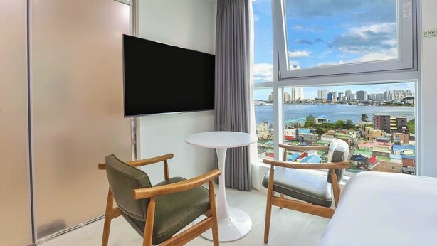 A hotel room with a large window offering a city view, a flat-screen TV mounted on the wall, and two wooden chairs with a small round table. - Hotel BERITH (Sokcho)