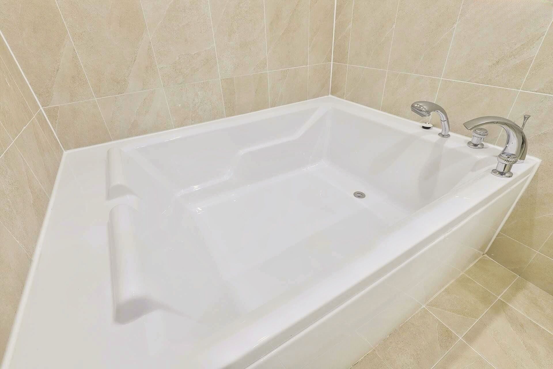 Una bañera blanca con dos llaves y desagüe en un baño con paredes de azulejos color beige.