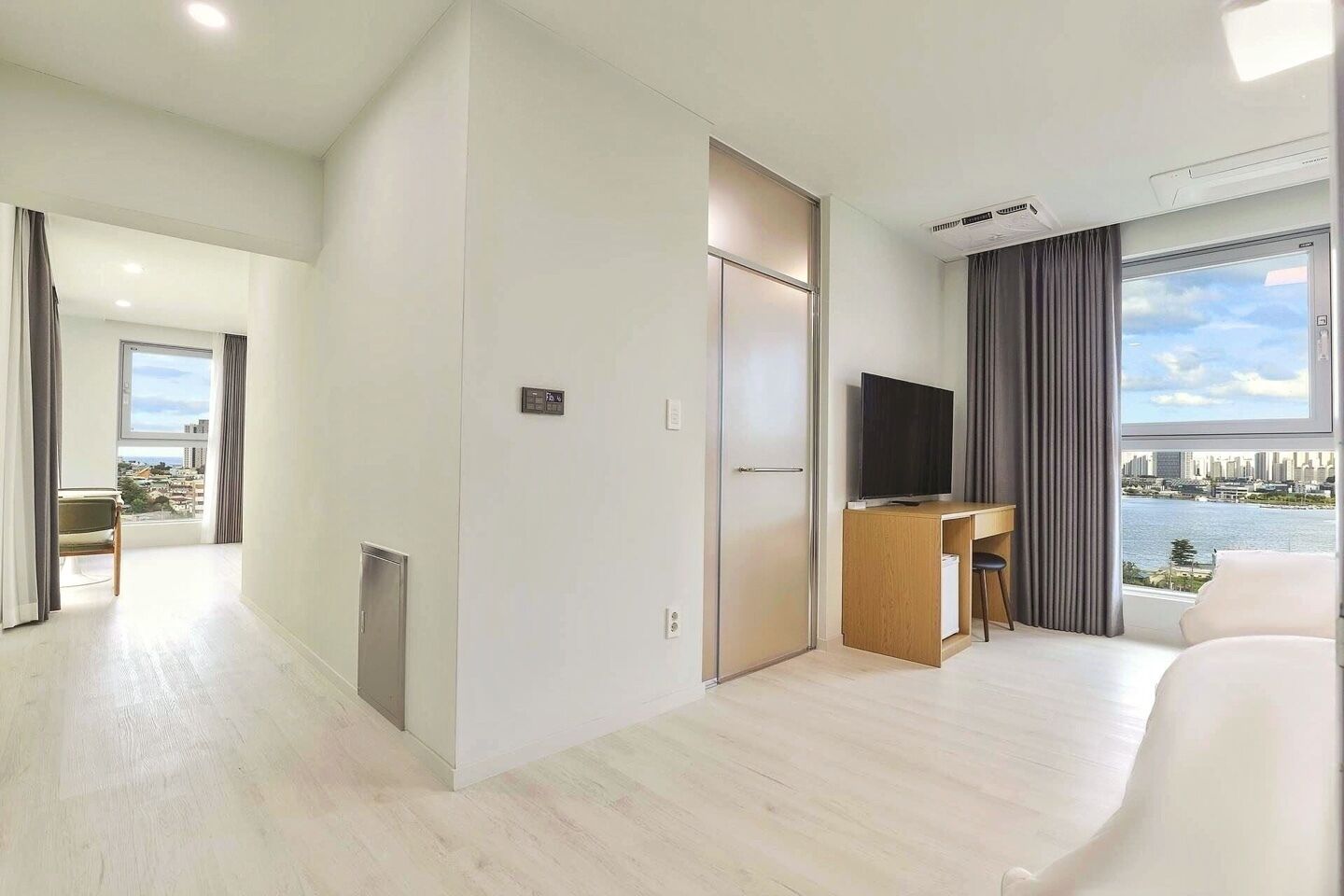 A modern hotel room with a city view, a desk with a chair, a television, and a large bed.