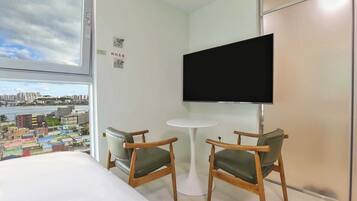 A hotel room with a bed, two chairs, a small table, a TV, and a view of a cityscape.