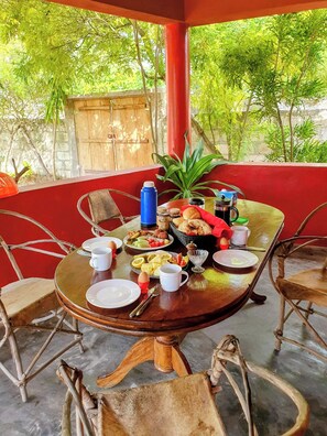 A round wooden table with chairs, a red wall, and a view of greenery.