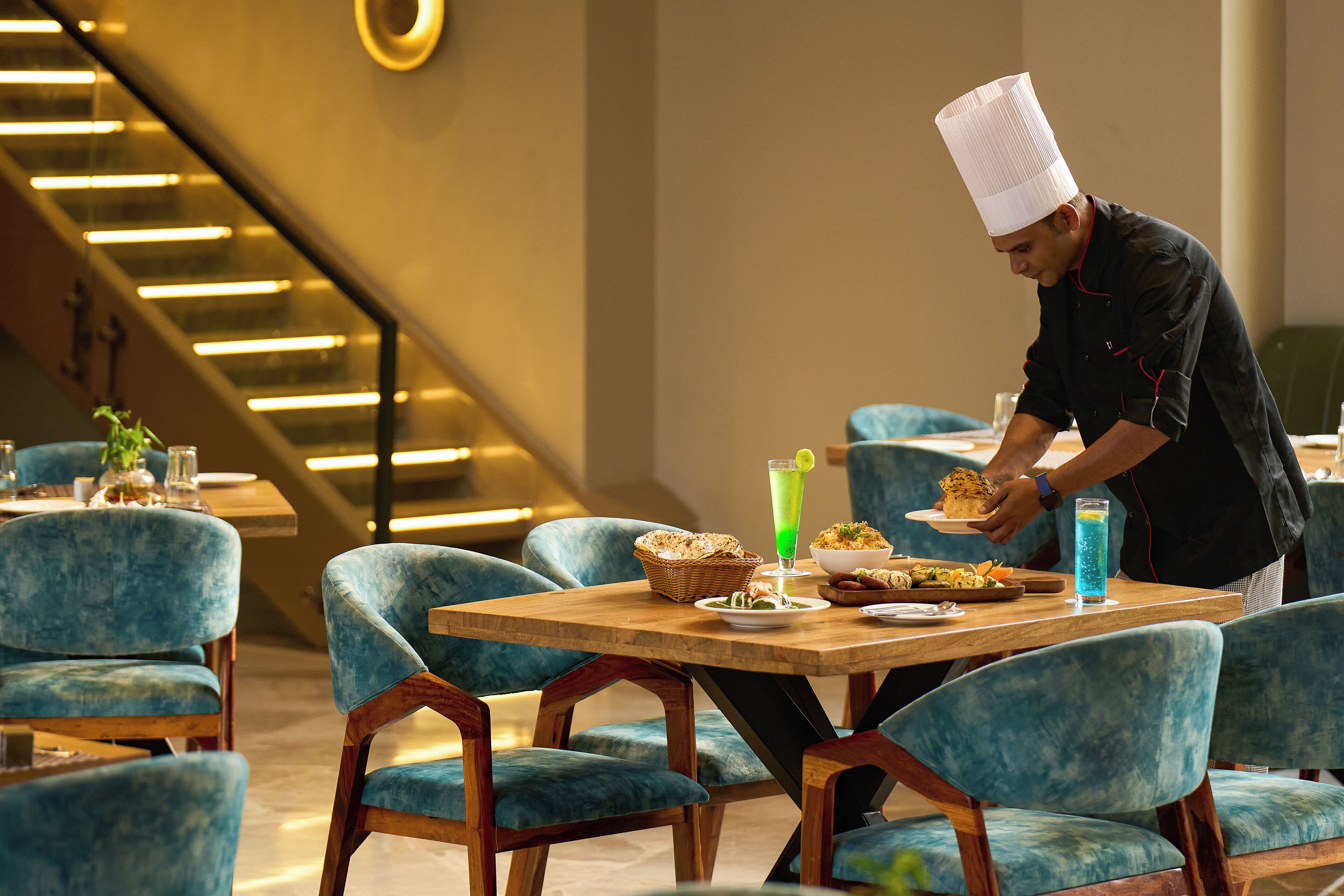 A restaurant with blue upholstered chairs, a wooden table, and a chef serving food.