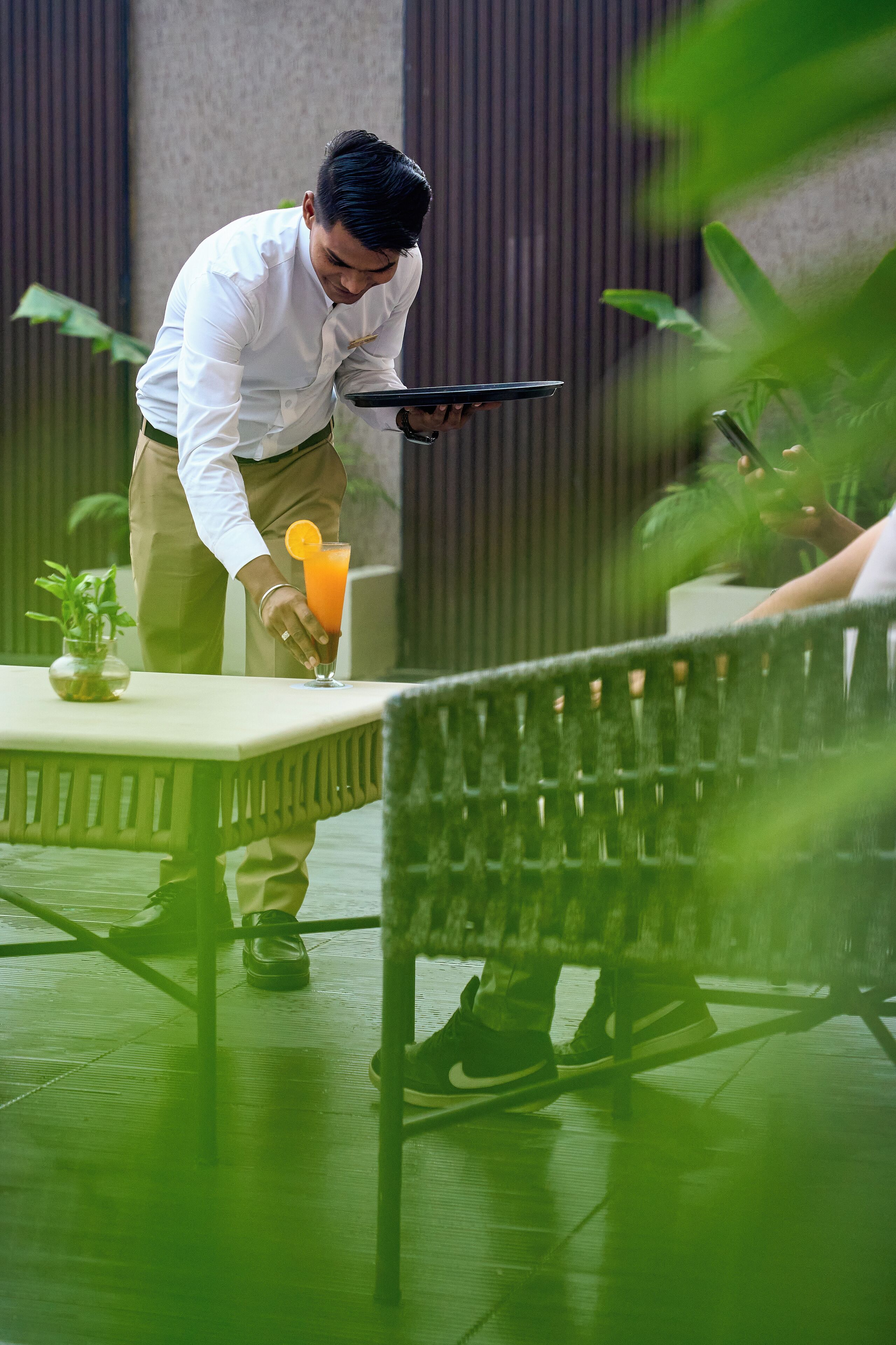 A waiter serving a drink to a guest at an outdoor table with a green chair.